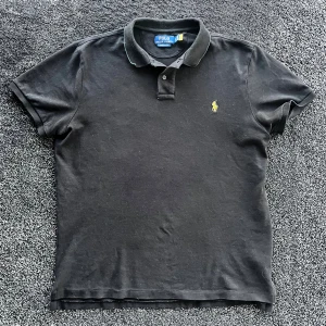 Polo Ralph Lauren pike - Svart pikétröja från Polo Ralph Lauren med klassisk krage och två knappar framtill. Tröjan har gul broderad logga på bröstet och är gjord i bomull. Passformen är slim och modellen har korta ärmar. Skick 8/10, storlek L.                              Pris går att diskutera✅
