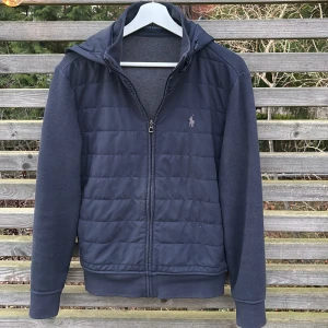 Mörkblå cardigan från Polo Ralph Lauren - Snygg mörkblå cardigan från Polo Ralph Lauren med quiltad framsida och klassisk logga på bröstet. Tröjan har huva, dragkedja och långa stickade ärmar. Mycket bra skick 8,5/10. Nypris 2999. Priset kan diskuteras vid snabb affär. 