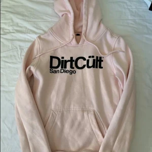 Ljusrosa DirtCült hoodie med tryck - Säljer en ljusrosa hoodie från DirtCült med stor svart logga och texten San Diego på bröstet. Tröjan har huva med snörning och en klassisk magficka. Perfekt för dig som gillar streetwear och vill ha en chill look.