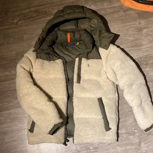 Teddyjacka från Polo Ralph Lauren - Snygg teddyjacka från Polo Ralph Lauren i beige och olivgrönt. Jackan har mysigt teddyfoder, gröna detaljer och dragkedjor, samt huva och orange snörning som kommer till ifall man vill sätta på dom på dragkedjan. Perfekt för kalla dagar och har en modern, loose passform.
