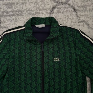 Grön mönstrad vindjacka från Lacoste - Säljer en grön vindjacka från Lacoste med geometriskt mönster och klassisk krokodillogga på bröstet. Jackan har svarta och vita ränder längs axlarna, dragkedja framtill och ribbade muddar. Regular fit och sportig vibe, perfekt för dig som gillar streetstyle.