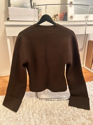 Brun kofta från Gina Tricot - Snygg brun kofta från Gina Tricot i storlek S. Koftan har v-ringad hals, två knappar framtill och långa ärmar med breda muddar och knappdetalj. Materialet känns mjukt och är stickat, perfekt för lager på lager under höst och vinter.