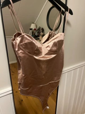 Satin body - Säljer en champagnefärgad  från NA-KD i glansig satin med smala axelband och markerade kupor. Bodyn har en figurnära passform och snygga sömdetaljer, perfekt för fest!