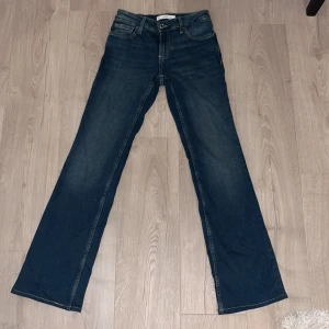 Blå bootcut jeans från MNG, stl 34 - Snygga mörkblå jeans från MNG i storlek 34 med klassisk bootcut passform. Jeansen har fem fickor, gylf med dragkedja och knapp, samt kontrastsömmar. Perfekta för dig som gillar en retro vibe och vill ha ett par jeans med lite utsvängda ben.