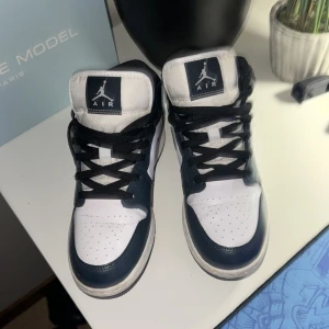 Nike Air Jordan 1 Mid marinblå/vit - Nike Air Jordan 1 Mid sneakers i marinblått och vitt med klassisk svart swoosh och snörning. Skorna har hög siluett, perforerad tå och robust gummisula. Materialet är skinn och textil, med svart foder och vit Air Jordan-logga på sidan. Perfekt för dig som gillar streetstyle. Storlek 40