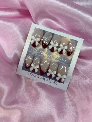 WhiteBows (S);Medium Almond - Säljer ett set handgjorda press-on naglar i S: 15 mm, 11,5 mm, 12 mm, 11 mm, 9 mm. Naglarna har en nude bas, vinröda tippar och 3D vita rosetter. Formen är medium längd i almondform och varje nagel har små pärlhjärtan. Perfekt för dig som vill ha en söt och unik look.