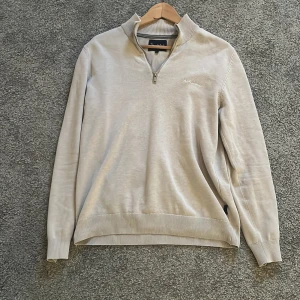 Beige half zip tröja från Bondelid - Snygg beige långärmad tröja från Bondelid med ribbad krage och muddar. Tröjan har en stilren half zip-dragkedja vid halsen och diskret broderad logga på bröstet. Perfekt för dig som gillar klassisk och clean stil. Tröjan är i storlek S men passar även XS.