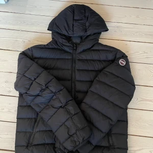 Colmar jacka - Svart dunjacka från Colmar med huva och dragkedja framtill. Jackan har quiltad design, två sidofickor med dragkedja och en rund logotyp på ärmen. Jackan är i bra skick men med lite skada på baksidan av jackans resårband som inte syns när du använder den. Pris kan diskuteras vid snabb affär 
