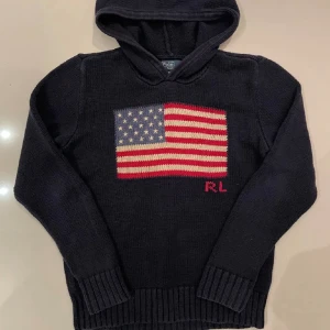 Mörkblå stickad hoodie från Polo Ralph Lauren - Snygg mörkblå stickad hoodie från Polo Ralph Lauren med amerikansk flagga på bröstet och RL-broderi i rött. Tröjan har huva, långa ärmar och breda ribbade muddar vid ärmslut och nederkant. Perfekt för dig som gillar klassisk sthlmstil.