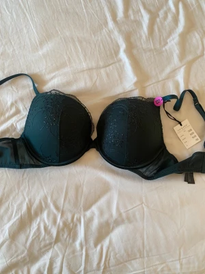 Grön bh från Primark - Supersnygg mörkgrön bh från Primark med broderade spetsdetaljer och lätt glansig yta. Axelbanden är justerbara och kuporna har ett elegant mönster. Push up bh med vaddering som är mjuk. Helt ny.  Storlek 80B europeisk storlek
