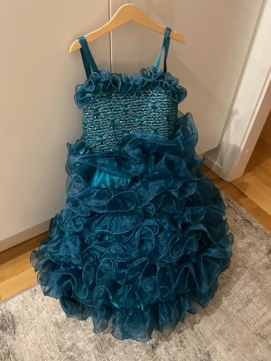 Turkos långklänning med volanger - En riktigt maffig turkos långklänning med tunna axelband och massor av volanger. Livet har ett broderat blommönster och volangkant upptill. Kjolen är superfluffig med lager på lager av transparent tyg som ger en dramatisk look.