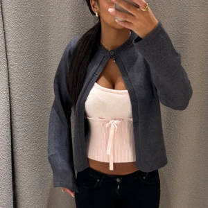 Mörkgrå kofta med knapp - Säljer en stilren mörkgrå kofta med rund hals och en enkel knapp framtill. Koftan har långärmade, lite loose ärmar och är tillverkad i mjukt stickat material. 