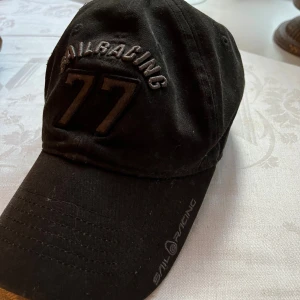 SailRacing keps svart - Svart keps från Sail Racing med broderad text och siffran 77 framtill. Justerbar rem baktill med metallspänne och Sail Racing-logga. Klassisk böjd skärm och stilren design, perfekt för en sportig look.