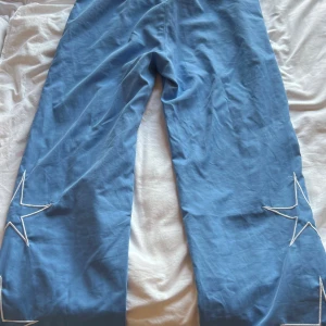 Blå campus täckbyxor med stjärnor - Säljer ett par ljusblå täckbyxor från Campus med vita detaljer och broderade stjärnor längs benen. Byxorna har elastisk midja med svart snörning, vita passpoaler längs sidorna och fickor. Perfekta för dig som gillar unik stil och coola detaljer.