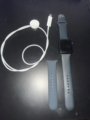 Apple Watch SE 44mm GPS Aluminium (Midnatt färg) - Säljer en Apple Watch med svart silikonarmband och extra armbandsdel. Klockan har en fyrkantig skärm och kommer med laddkabel. Perfekt för dig som vill ha koll på mobilen direkt på handleden och gillar stilren tech.