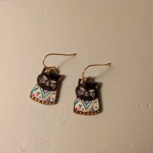Örhängen med svarta kattmotiv - Unika örhängen med svarta katter och färgglada mönster på kroppen. Hängena har guldiga krokar och kattens kropp är dekorerad i blått, rosa och vitt med grafiska detaljer. Perfekt accessoar för dig som älskar katter och vill sticka ut.