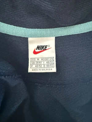 Nike vindjacka i mörkblå och röd - Nike vindjacka i mörkblått med en bred röd panel över bröstet och klassisk vit swoosh-logga. Jackan har dragkedja framtill, hög krage och långärmade ärmar. Perfekt för dig som gillar sportig och chill stil.