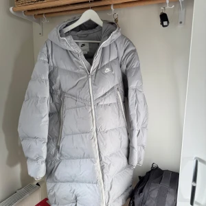 Grå Nike pufferjacka med huva XL - Säljer en grå pufferjacka från Nike i storlek XL med huva och Storm-Fit teknologi. Jackan har dragkedja framtill, två sidofickor med dragkedja och ett stort Nike-logga på bröstet. Perfekt för kalla dagar och har en clean, sportig vibe. Har ett hål på insidan av kragen till dragkedjan. Det syns på sista bilden. 