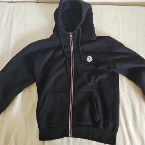 Svart Moncler hoodie med dragkedja - Svart hoodie från Moncler med broderad logga på bröstet och ärmen. Tröjan har huva med snörning, hel dragkedja med röd, vit och blå detalj, samt två fickor framtill. Tillverkad i mjuk bomull för skön komfort. Perfekt för dig som gillar stilrena märkesplagg. Har defekter! Därav pris.