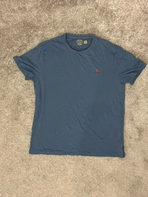 Blå t-shirt från Polo Ralph Lauren - Snygg blå t-shirt från Polo Ralph Lauren med klassisk röd logga broderad på bröstet. Modellen är kortärmad och har en rund halsringning. Tillverkad i mjuk bomull och har en clean, stilren look som funkar till det mesta. Använd tröja så slitage finns