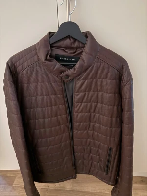 Brun quiltad skinnjacka Zara Man - Snygg brun quiltad skinnjacka från Zara Man med ståkrage och dragkedja framtill. Jackan har horisontella sömmar, två fickor med dragkedja och en stilren look som passar perfekt till höst och vinter. Materialet är skinn och jackan har långärmade ärmar.