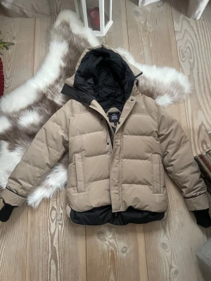 Beige pufferjacka från Canada Goose - Säljer en beige pufferjacka från Canada Goose med svart foder och huva. Jackan har ribbade muddar, dragkedja och knappar framtill samt två stora fickor. Knappt använd så den är i perfekt skick! Tror att det är en herrmodell men jag använde den som en oversized jacka.