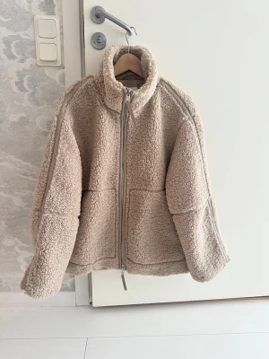 Beige pilejacka med dragkedja, slutsåld! - Mysig beige teddyjacka med hög krage och grov dragkedja framtill. Jackan har två stora fickor och en loose passform som ger en chill vibe. Perfekt för kalla dagar och enkel att slänga på över en hoodie. Materialet är fluffigt fleece och ger en skön känsla. Jackan är slutsåld.