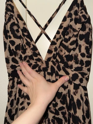 Leopardmönstrad långklänning - Säljer en snygg långklänning med leopardmönster i beige och svart. Klänningen har djup v-ringning och smala axelband som korsas i ryggen. Materialet känns mjukt och följsamt, perfekt för dig som gillar att sticka ut med djurprint.