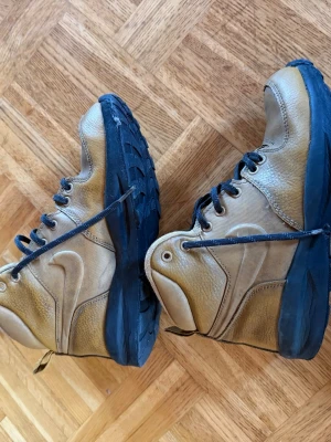 Nike boots i beige och svart, strl 37.5 - Säljer ett par robusta Nike boots i beige med svarta detaljer och snörning. Skorna har en rund tå, platt sula och är tillverkade i skinn och syntetmaterial. Perfekta för dig som gillar outdoor och vill ha en cool stil med klassisk Nike-logga på sidan.