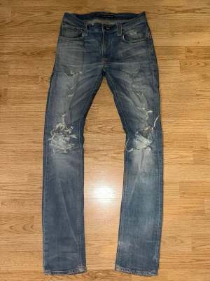 Blå slitna jeans från Nudie Jeans - Säljer ett par blå jeans från Nudie Jeans med slitna detaljer och hål på benen. Jeansen har klassisk femficksdesign, normal passform och är tillverkade i bomull. Perfekta för en avslappnad och trendig look.