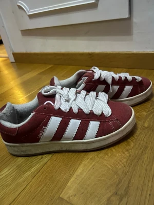Adidas Campus röda sneakers - Säljer ett par Adidas Campus sneakers i rött med klassiska vita ränder på sidorna och vit sula. Skorna har rund tå, platt sula och snörning med breda vita skosnören. Ovandelen är i mocka och insidan är ljusgrå. Perfekt för dig som gillar retrostil och streetwear.