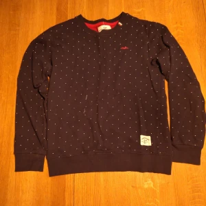 Mörkblå prickig tröja Hampton Republic - Snygg mörkblå tröja från Hampton Republic med små vita prickar över hela. Rund hals och långa ärmar, insida i röd mesh. Tillverkad i 80% bomull och 20% polyester, perfekt för dig som gillar klassisk stil med en twist.