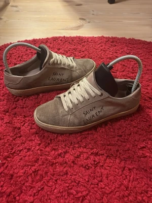 Saint Laurent beige sneakers i mocka - Snygga beige sneakers från Saint Laurent med handskriven logga på sidan. Skorna har rund tå, platt sula och snörning framtill. Tillverkade i mjuk mocka med detaljer i skinn och gummisula. Perfekta för dig som gillar stilrena och exklusiva sneakers. Hör av dig vid minsta fundering och jag fraktar alltid en till två dagar efter beställningen!