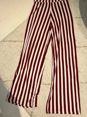 Randiga wide byxor i vinrött och vitt - Säljer ett par riktigt snygga wide byxor med vertikala ränder i vinrött och vitt. De har hög midja och är gjorda i ett mjukt bomullsmaterial som känns skönt mot huden. Perfekta för dig som gillar att sticka ut med coola mönster och avslappnad passform.