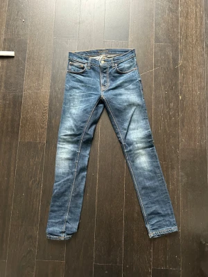 Blå jeans från Nudie Jeans Co, 28/32 - Snygga blå jeans från Nudie Jeans Co i storlek 28/32. Modellen har klassisk femficksdesign, orangea kontrastsömmar och coola slitningar framtill. Materialet är slitstark bomull och passformen är slim med raka ben. Bakfickorna har Nudie Jeans signaturbroderi.