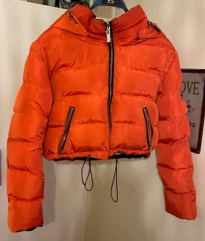 Svart och orange pufferjacka Manière De Voir - Säljer en croppad pufferjacka från  Manière De Voirmed fet huva och snörning i nederkant. Jackan är vändbar med svart utsida och knallig orange insida, har dragkedja framtill och två fickor med zip. Perfekt för dig som vill sticka ut i vinter. Kan användas både som svart och orange sidan.