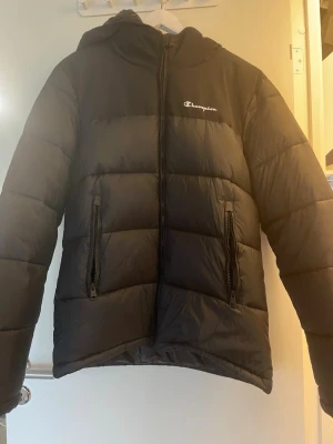 Svart pufferjacka från Champion - Svart pufferjacka från Champion med huva och två fickor med dragkedja. Jackan har ett quiltat mönster och logga på bröstet samt insidan. Perfekt för kalla dagar och har en riktigt chill vibe.