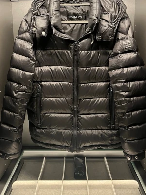 Svart pufferjacka från BVALDi - Säljer en svart pufferjacka från BVALDI med huva och dragkedja framtill. Jackan har en clean look med diskret logga på ärmen och två sidofickor med dragkedja. Perfekt för slutet på vintern och början på våren då den håller värmen bra samtidigt som den andas. Jackan är i fint skick utan några defekter. Vid frågor och funderingar är det bara att höra av sig i PM 🤝🫡😀