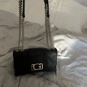 Guess väska - Handväska från guess, svart. Riktigt snygg o passar till allt. G:et på framsidan har blivit litee rostigt men annars jättefint skick. Handtaget o kedjorna är helt rostfria och lädret är i jättebra skick.