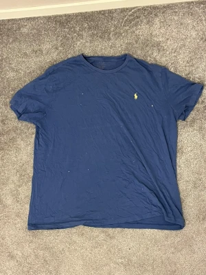 Blå t-shirt från Polo Ralph Lauren - Snygg marinblå t-shirt från Polo Ralph Lauren med klassisk gul logga på bröstet. Modellen är kortärmad och har en rund halsringning. Tillverkad i mjuk bomull som är skön mot huden. Perfekt för dig som gillar stilrena och enkla plagg med ikonisk branding. Tröjan är använd så slitage finns 