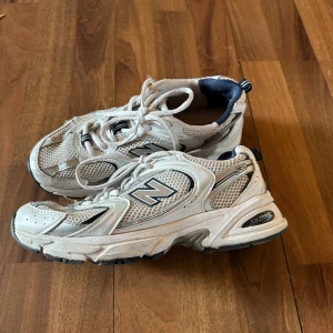Vita New Balance 530 sneakers - Säljer ett par klassiska New Balance 530 sneakers i vitt med grå och mörkblå detaljer. Skorna har meshpaneler för extra andningsförmåga, snörning och en rejäl sula med ABZORB-dämpning. Välanvända men fortfarande snygga på