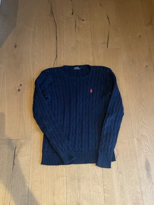 Mörkblå stickad tröja Polo Ralph Lauren - Säljer en mörkblå stickad tröja från Polo Ralph Lauren i storlek s . Tröjan har kabelstickat mönster, rund hals och en röd broderad logga på bröstet. Tillverkad i 100% bomull och har långa ärmar med ribbade muddar. Perfekt för dig som gillar klassisk stil.