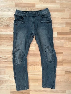 Grå jeans från Balmain med dragkedjor storlek 32 - Säljer ett par grå jeans från Balmain Paris med coola detaljer som förstärkta knän, dragkedjor vid bensluten och fickor med zip. Jeansen har en snygg ribbad panel bak och markerade sömmar som ger en edgy vibe. Perfekt för dig som gillar streetstyle och vill sticka ut.