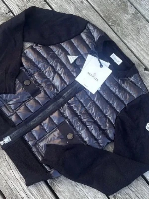 Svart quiltad pufferjacka från Moncler - Säljer en svart pufferjacka från Moncler med quiltad framsida i glansigt material och stickade ärmar. Jackan har rund hals, två fickor med knapp och dragkedja framtill. Perfekt för dig som vill ha en snygg och stilren jacka med exklusiv känsla.