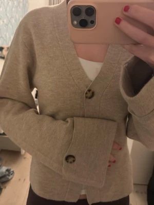 Beige kofta från Gina Tricot - Säljer en beige kofta från Gina Tricot med två stora, mönstrade knappar framtill. Koftan har långärmad design och är tillverkad i mjukt stickat material. Perfekt att slänga på sig när det är lite kyligt ute. Snygg och enkel att matcha med olika outfits.