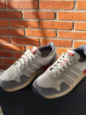 Adidas retro sneakers i grå och röd 36 2/3 - Säljer ett par klassiska Adidas sneakers i retrostil med ovandel i grått och vitt tyg, detaljer i mocka och röd accent på hälen. Skorna har snörning, rund tå och den ikoniska Adidas-loggan på baksidan. Perfekta för dig som gillar old school-vibbar.