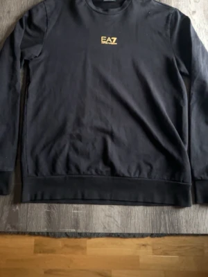 Svart sweatshirt från EA7 Emporio Armani - Svart långärmad sweatshirt från EA7 Emporio Armani med gul logotyp på bröstet och gul detalj i nacken. Tröjan har rund hals och ribbade muddar vid ärmslut och nederkant. Tillverkad i mjuk bomull som är skön att ha på sig.