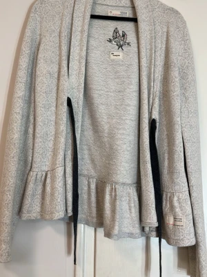 grå kofta  - Säljer en ljusgrå kofta från odd molly med subtilt mönster och volangdetalj nedtill. Koftan har långa ärmar och knytband framtill. Perfekt för lager-på-lager och har en mjuk bomullskänsla. Broderad detalj vid nacken ger extra stil.