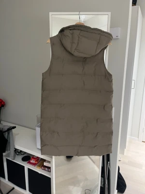 Beige lång dunväst från Zara - Säljer en beige, lång dunväst från Zara med huva och dragkedja framtill. Västen har två fickor med dragkedja och snörning vid huvan. Perfekt för lager-på-lager och chill stil under kyliga dagar.