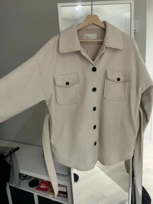Beige overshirt från NLY Trend - Snygg beige overshirt från NLY Trend med stora svarta knappar och två bröstfickor. Jackan har ett bälte i midjan och är tillverkad i mjukt material som ger en chill vibe. Perfekt för lager-på-lager och har en loose passform med långärmade ärmar.